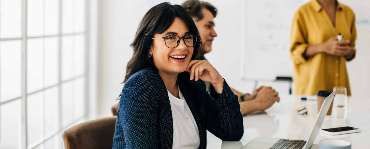 A person smiling with glasses sat at a laptop during a board meeting.