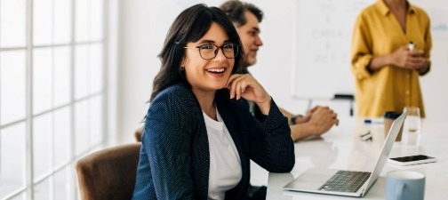 A person smiling with glasses sat at a laptop during a board meeting