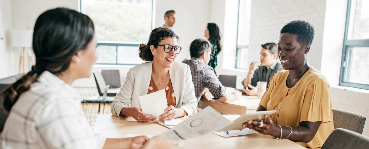 Three businesswomen sat at a table, smiling and discussing a report in a brightly lit office environment