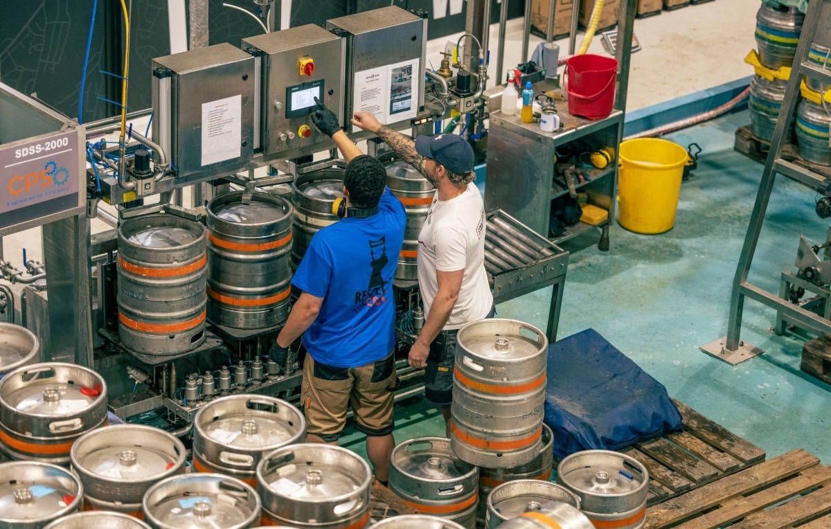Two brewery workers operating a keg-filling machine inside a brewery, surrounded by stacked metal beer kegs and industrial equipment.