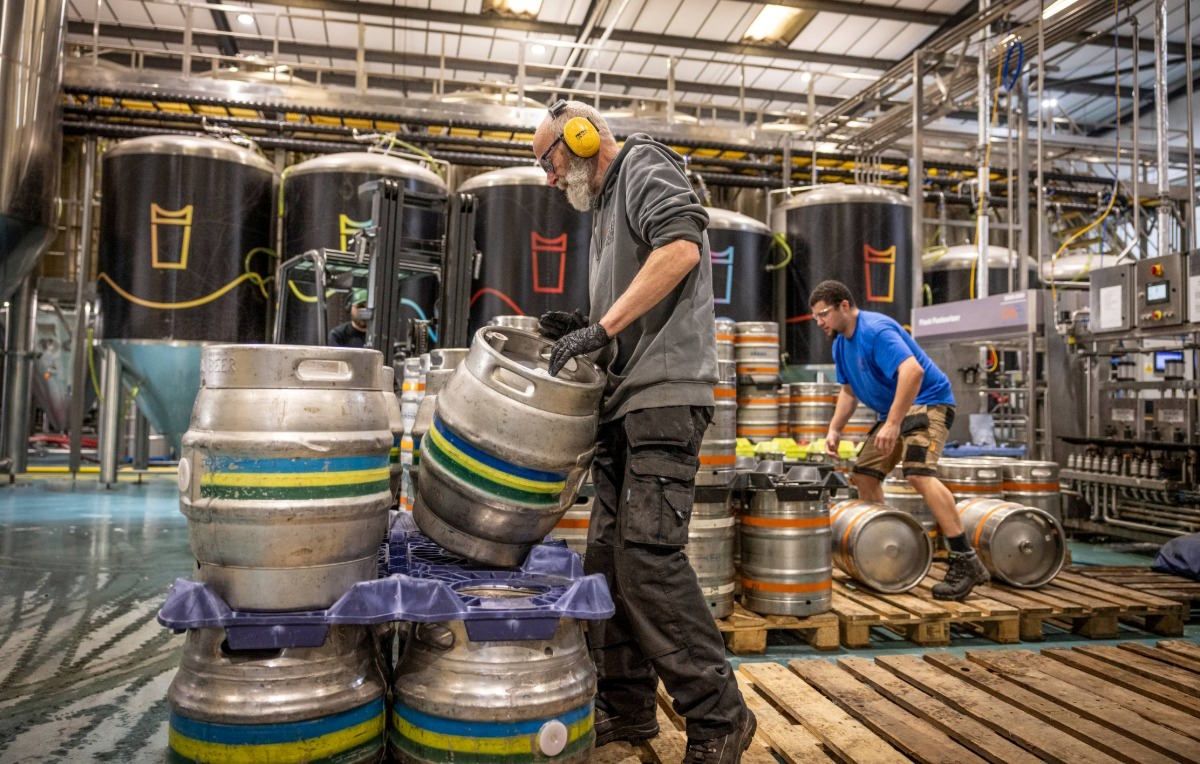 Brewery workers lifting and moving metal kegs on pallets inside a large production space with fermentation tanks in the background.