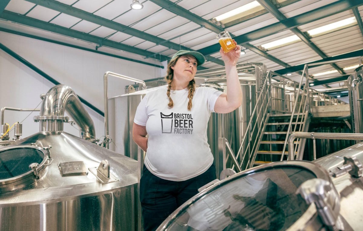 Bristol Beer Factory worker inspecting a glass of beer while standing among stainless steel brewing tanks in a production facility.