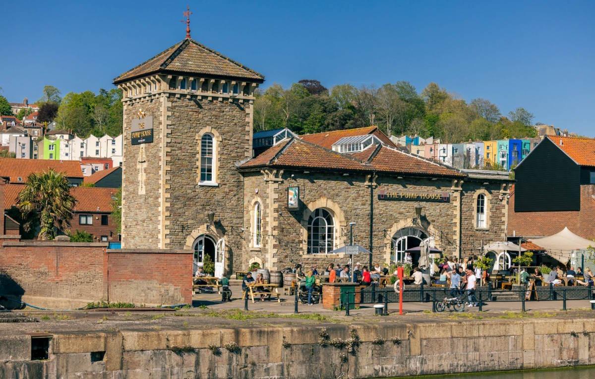 Historic stone building on Bristol’s harbourside with outdoor seating area, people gathered outside The Pump House pub on a sunny day, with colourful houses on the hillside behind.