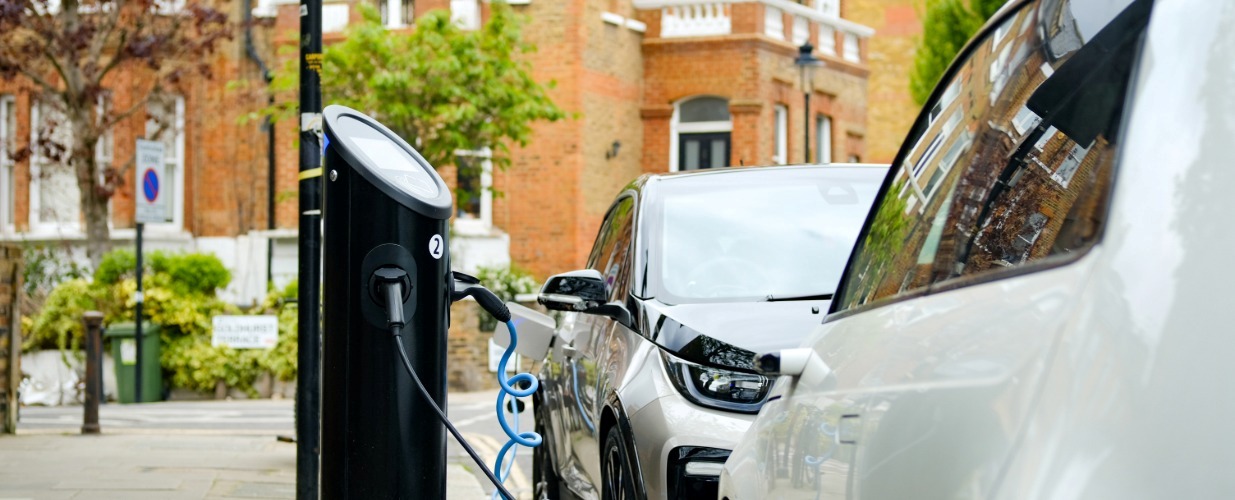 Two electric cars charging on a residential street with buildings and greenery in the background.