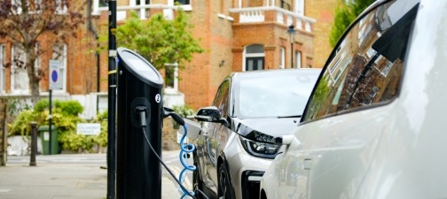 Two electric cars charging on a residential street with buildings and greenery in the background.