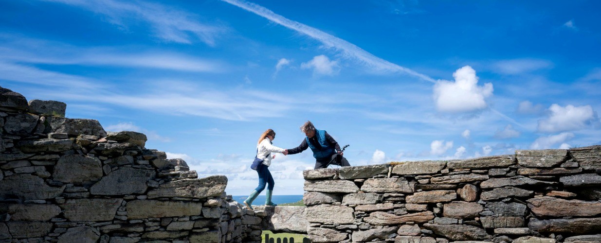 Two people help each other across a gap between stone walls under a blue sky.