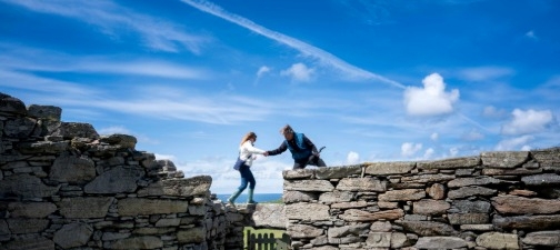 Two people help each other across a gap between stone walls under a blue sky.