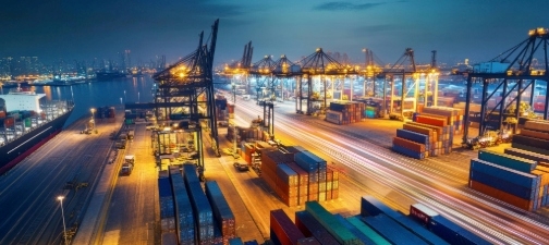 Aerial view of an illuminated container port at dusk, showing cranes and stacked shipping containers in operation