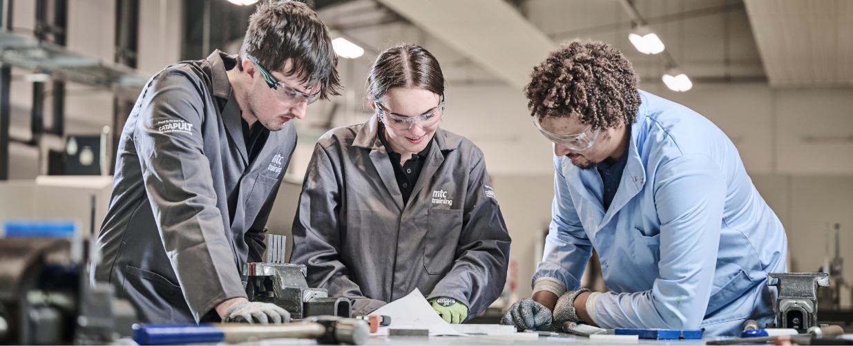 Two apprentices at the mtc around a workbench with a teacher