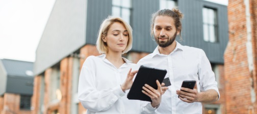 Two people in white shirts stand outside a modern building, one holding a tablet, the other a smartphone.