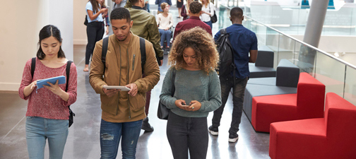 Three students walk through campus using their mobile phones