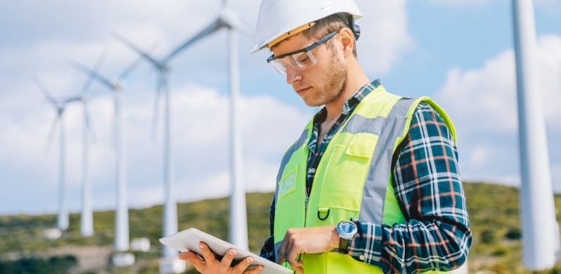 Man in High-Visibility Jacket looking at computer tablet with Wind Turbines behind him