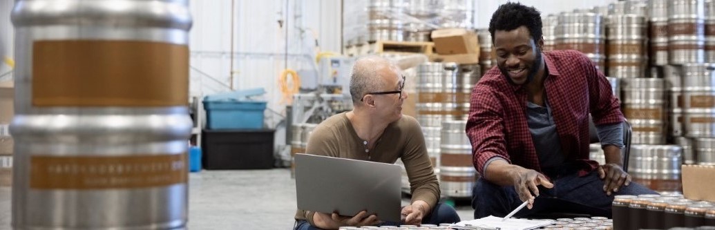 Two workers in a drinks warehouse with a laptop