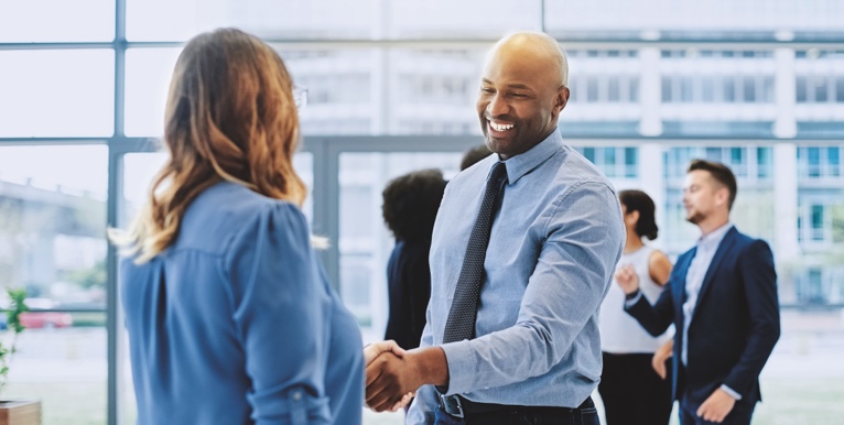 Two people smile and shake hands in a busy office.