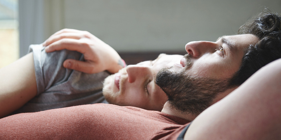 Male couple lying down, one of the men is asleep the other is gazing into the distance.