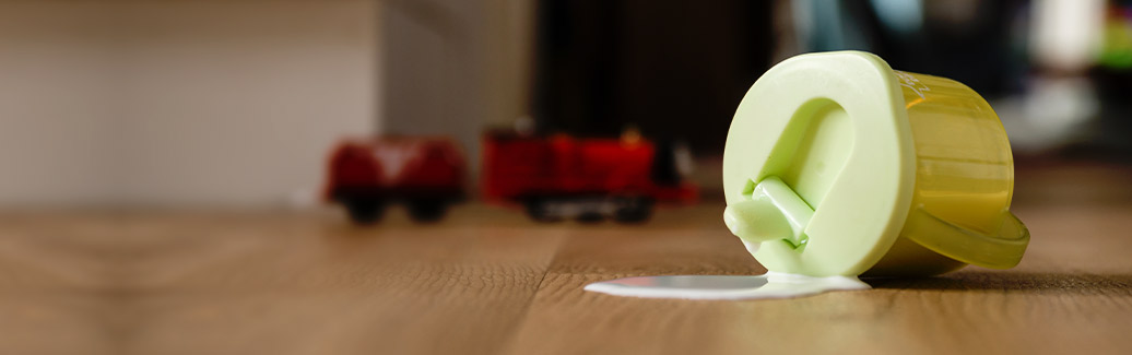 A child's sippy cup on it's side, with milk dripping onto the floor.