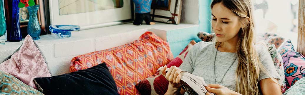 Woman sat surrounded by cushions reading a book.