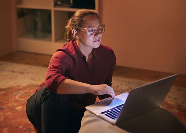 Young woman sat on the floor of her dimly lite living room floor as she looks at her laptop.