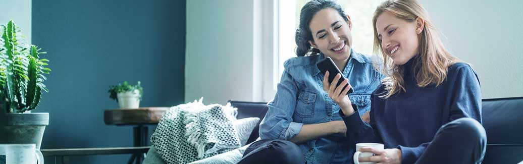 Two women sat on the sofa, smiling at their mobile phone.